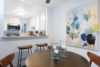 round table with 4 wooden chairs, colorful painting on the wall with breakfast bar and kitchen in the background.